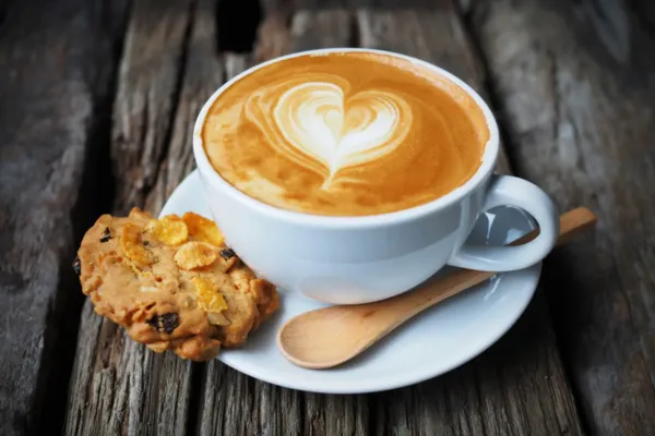 Taza blanca de café con arte latte en forma de corazón, acompañada de una galleta y una cuchara de madera sobre una superficie de madera rústica.